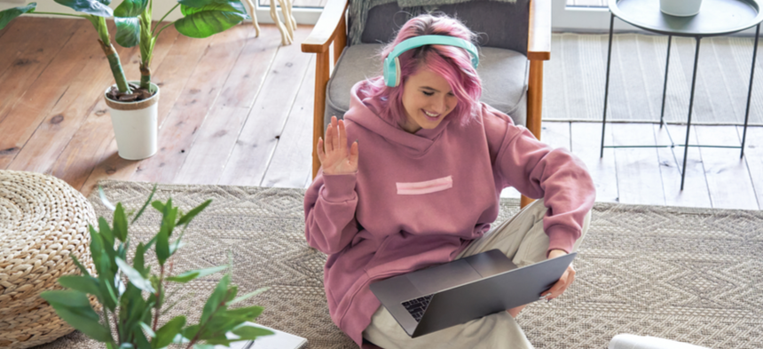 Teen wearing pink hoodie and teal headphones, sitting on floor, waving to friends on laptop